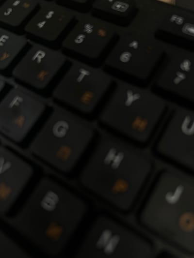 The image is a close-up, low-angle, blurred photograph of a black keyboard. The keys display white letters and numbers, with some keys also having orange symbols. The lighting is dim, casting shadows and creating a dark, moody atmosphere. There is no indication of the city of Yoshkar-Ola, Russia, or any other geographical location. The image focuses solely on the keyboard, with no people, actions, or interactions depicted. The environment is confined to the surface of the keyboard itself.