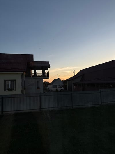 The image depicts a rural scene in Chemerne, Ukraine, during sunset. The sky is a gradient of soft blues and oranges, indicating the time of day. In the foreground, several buildings are visible. The closest building is a two-story house with a brown tiled roof and a yellow facade, featuring white-trimmed windows. To its left and slightly behind, another building with a similar brown roof is partially visible. Further back, a multi-story structure with an open, lattice-like second story stands. Behind this, a white van is parked. The buildings are separated by a grey, corrugated metal fence. To the right of the fence, a dense area of dark green grass or foliage dominates the scene. The overall atmosphere suggests a peaceful evening in a residential area. There is no visible text in the image.