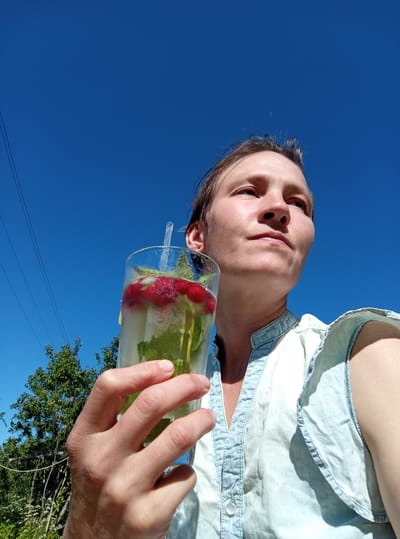 The media file shows a person holding a glass of iced beverage outdoors against a clear blue sky. The person, who appears to be female, is wearing a light blue denim shirt. The glass contains ice, mint leaves, raspberries, and a slice of lemon or lime. A clear plastic straw is visible in the glass. The hand holding the glass is positioned in the upper left of the frame. In the background, out of focus, are green trees and power lines. The image is taken from a low angle, looking upwards. The bright sunlight suggests it is daytime, likely a warm and sunny day. The overall mood seems relaxed and summery. No text is visible in the image. The location context is Ostrovtsy, Russia.
