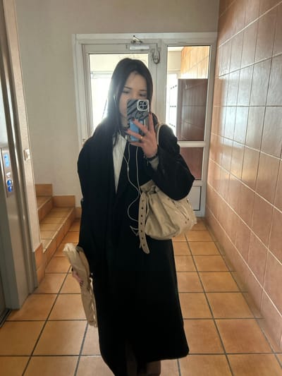 An individual, appearing to be female with long dark hair, captures a mirror selfie in the hallway of a building located in Cachan, France. She is dressed in a dark, long overcoat worn over a lighter garment, with white wired earbuds visible. Her left hand, adorned with rings and dark nail polish, holds a smartphone featuring a distinctive patterned case, positioned to photograph her reflection. A light-colored, studded drawstring shoulder bag hangs over her right shoulder. Her right hand grasps a brown paper bag, consistent with packaging for an elongated item such as a baguette. The immediate environment consists of uniform brown square tiles covering both the floor and the wall to the right. The left wall is a light, untextured surface where a vertical mirror is mounted, along with an illuminated elevator call panel. Behind the individual, a white-framed door with clear glass inserts leads to an adjacent space, which also features tiled surfaces. A tiled staircase ascends on the far left side of the frame, beyond the elevator panel.