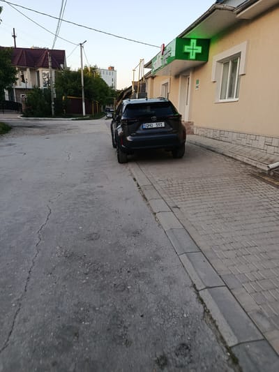 The image depicts a street scene in Bălți, Moldova, likely in a commercial or mixed-use area. In the foreground, a dark-colored SUV is parked on the right side of the street, facing away from the camera. The license plate is visible and reads "QHO 591". The street itself is paved with asphalt, showing signs of wear and cracking, with a distinct sidewalk made of interlocking pavers running along the right side of the road. Adjacent to the sidewalk is a building with a light-colored facade, featuring a prominent green illuminated cross sign, indicative of a pharmacy. The sky is clear and appears to be late afternoon or early evening, with soft lighting casting gentle shadows. In the background, residential buildings and trees are visible, suggesting a semi-urban environment. The overall atmosphere is calm and ordinary, with no discernible activity or people present in the immediate vicinity of the vehicle.