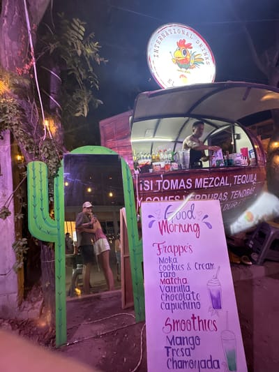 The image depicts a nighttime scene in what appears to be Brisas de Zicatela, Mexico, showcasing a mobile beverage cart. The cart is adorned with the words "ISI TOMAS MEZCAL, TEQUILA." A man stands inside the cart, seemingly preparing drinks. A sign next to the cart advertises "Good Morning" beverages, listing options like Frappe's (Moka, Cookies & Cream, Taro, Matcha, Vanilla, Chocolate, Capuchino) and Smoothies (Mango, Fresa, Chamonada). A cactus-shaped photo frame is positioned nearby, reflecting an image of people in the frame. An illuminated sign reading "International Drinks EL" is visible above the cart. The surrounding environment includes plants and what appears to be a dimly lit outdoor area.