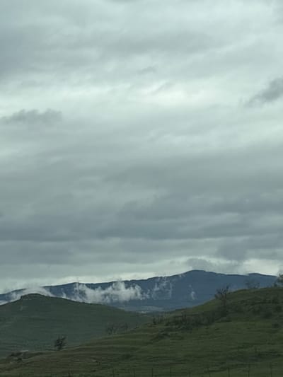 The image shows a view of a mountain range partially obscured by clouds. The right side of the image depicts a green, grassy hillside with sparse trees. The mountain itself is visible in the center, a darker blue color. A dense cloud cover dominates the left portion of the image.
There is no information given that the location is Levunovo, Bulgaria and the image shows nothing that indicates this.