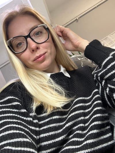 A light-skinned female subject with long, straight blonde hair and light-colored eyes is depicted in an indoor setting. She wears black-framed glasses with white accents, a white collared shirt, and a black knitted sweater featuring horizontal white stripes. Her left hand is partially visible, with fingers touching her hair near the right temple. The subject's expression is neutral with a subtle upward curve of the lips. The background is out of focus, showing a light-colored wall or surface and indistinct objects, including a blurry green item potentially covered in clear plastic in the mid-right. No verifiable visual information is present within the image to confirm the specified location of Voskresensk, Russia.