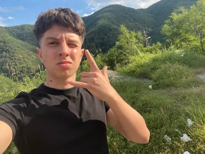 The image is a selfie taken outdoors in a rural, mountainous environment. A young person with dark, messy hair is centered in the frame, wearing a black t-shirt. They are winking and making a "shaka" hand gesture. The background consists of lush green hillsides covered in trees and vegetation, under a bright, partly cloudy sky. The overall impression is of a casual, outdoor activity in a natural setting, likely in a location like Pechinska, Bulgaria, known for its scenic mountainous terrain.
