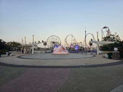 The image depicts an outdoor plaza in front of amusement park attractions at dusk. In the foreground, a circular paved area features distinct zones of gray and purple concrete, bordered by dark gray concrete strips with a textured pattern. A large, pale pink inflatable sculpture of a sleeping bear holding a guitar is centrally located in the plaza, surrounded by decorative lighting.
In the mid-ground, a white shaded structure with tall, ornate pillars provides shelter. Several people are visible walking and sitting under this structure and along a low wall. Beyond this, the backdrop is dominated by amusement park rides, including a Ferris wheel on the left and several roller coasters with red tracks and white supports. Illuminated circular signs are visible on the roller coaster structures.
The background shows more of the amusement park structures and some trees and buildings. The sky is a clear, pale blue, indicating early evening or late afternoon. The overall scene suggests a recreational area, likely a theme park or amusement park, situated in the city of Li'an, China.