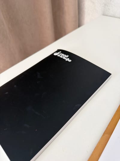 This media file shows a black notebook with white text on its cover, placed on a white surface that appears to be a table or desk. The notebook is angled, with its spine facing upwards and to the right. The text on the cover is in Cyrillic script and appears to be a logo or title, possibly related to a brand or publication. The background is blurred, suggesting a shallow depth of field, but hints of a wooden surface can be seen below the white surface. A thin wooden object, possibly a ruler or a piece of furniture, runs diagonally across the upper right portion of the image. The lighting suggests an indoor setting, possibly during the day. There are some smudges and faint marks on the black cover of the notebook.