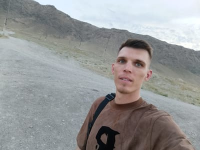 This is a selfie taken by a young man with short brown hair and blue eyes. He is wearing a brown t-shirt with a black graphic design and a black strap across his shoulder, suggesting he is carrying a bag. He has a light goatee and appears to be smiling slightly.
The background shows a mountainous, arid landscape with a gravelly path leading uphill. There are sparse patches of dry grass and several utility poles with wires scattered across the hillside. The sky is overcast with clouds, indicating a potentially mild or cool weather condition. The overall scene suggests an outdoor activity, possibly hiking or exploring in a remote, mountainous area. The lighting suggests it is either early morning or late afternoon.
The location is identified as Zarafshan City, Uzbekistan, which aligns with the arid, mountainous terrain. There is no visible text in the image. The man's expression is neutral to slightly pleasant, conveying a sense of calm or contentment.