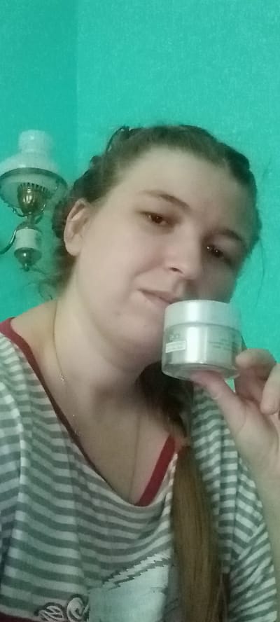 A woman, with her hair pulled back in a braid, holds a small, white jar of skincare product in her right hand, close to her face. She is looking towards the viewer, with a slight smile. She is wearing a grey and white striped t-shirt with a red trim around the neckline. A thin gold necklace with a small pendant is visible around her neck. In the background, to the left of the woman, is a vintage-style oil lamp mounted on the wall. The wall itself is painted a bright, turquoise color, consistent with the typical interior paint choices found in residential buildings in the city of Kitskany, Moldova.