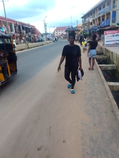The image depicts a street scene in Egbagu, Nigeria. The main subject is a young Black woman walking towards the viewer on a paved road. She is wearing a black t-shirt and black leggings, with blue and white flip-flops on her feet. She is carrying a dark item, possibly a bag or clothing, in her right hand. Behind her, another young Black woman is walking away from the viewer, wearing a black t-shirt and denim shorts, with flip-flops.
In the background, a yellow and black auto-rickshaw is parked on the left side of the road. Beyond the road, there are buildings lining the street. On the left, a red building with multiple floors and balconies is visible, featuring signs that read "OCE RENTALS." On the right, a taller, multi-story building has signs, one of which appears to be for "Gudnews C.A.cademy." Another sign on the right for "Jozam Tech" displays information about computer services. Various other people are seen walking along the street and sidewalks, and some are gathered near the buildings.
The scene appears to be during the day, with an overcast sky, suggesting cloudy weather. The overall atmosphere seems busy, typical of a commercial or residential street in a town. The lighting is diffused, and there are no strong shadows. The image captures everyday life on a street in Nigeria, with pedestrian and vehicular traffic, as well as commercial establishments.
