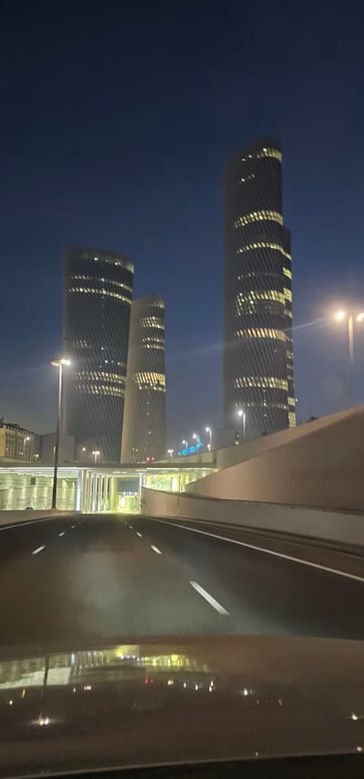 The image captures a nighttime view of the Lusail City skyline in Qatar, specifically featuring the distinctive architecture of the Lusail Towers and surrounding infrastructure. In the foreground, a dark asphalt road with white lane markings is visible, suggesting a driver's perspective, possibly from a vehicle. The road leads towards an illuminated underpass or tunnel structure.
Dominating the background are three tall, modern skyscrapers with unique, spiraling facades. The buildings are illuminated from within, with rows of yellow lights visible through the windows, indicating that they are occupied or lit for nighttime visibility. The tallest tower is on the right, with two slightly shorter, similarly designed towers to its left. The sky is a deep, dark blue, characteristic of nighttime.
Streetlights are strategically placed along the road and around the buildings, casting a warm glow. A concrete embankment or highway barrier curves along the right side of the road, separating it from the cityscape. In the distance, other less distinct buildings and city lights can be observed. The overall scene conveys a sense of urban development and modern architectural design within Lusail City. No people are visible in the image.