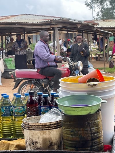 In Wang, Nigeria, an outdoor market scene unfolds under a cloudy sky. A man in a lavender jacket sits astride a red motorcycle, his hands on the handlebars. To his right, another man in a dark shirt holds a piece of food to his mouth. In the foreground, bottles of cooking oil and dark liquid are arranged on a table, with a large white bucket and a green basin behind them. Various stalls with corrugated metal roofs and wooden supports are visible in the background, where other people are browsing or working. The overall impression is one of a bustling, everyday market in a Nigerian city.