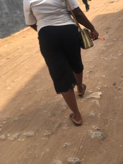 An adult individual walks away from the camera on an unpaved, reddish-brown dirt path in Egan, Nigeria. The individual wears a light-colored short-sleeved top, black knee-length culottes or a skirt with a patterned or lace hem, and dark-colored open-toed sandals. A gold-colored shoulder bag is worn across the body, with the right hand holding the bag's strap and a small red object. The ground is uneven, comprised of packed earth with scattered small rocks and patches of lighter-colored debris, displaying faint tire tracks. In the blurry background to the left, a grey concrete structure is visible, and another distant, blurry figure can be seen walking further up the path. The scene is illuminated by natural daylight.