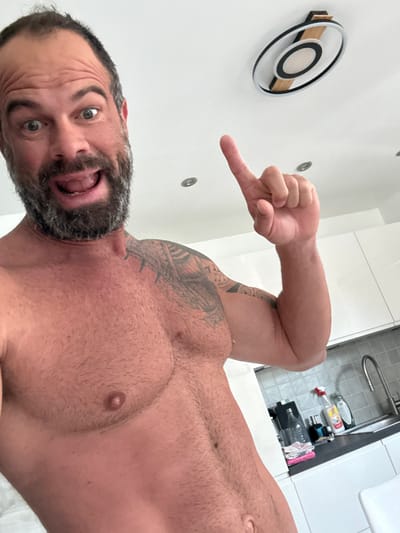 A shirtless man with a dark beard and visible arm tattoos takes a selfie from a low angle, looking up towards the camera. He has an expressive face with wide eyes and an open mouth, conveying a playful or surprised emotion. His right arm is bent, and his index finger is extended, pointing towards a decorative, circular object mounted on a white wall. This object features concentric rings in black, white, and a light wooden or beige finish.
The setting appears to be indoors, likely a bathroom or kitchen, suggested by the presence of a stainless steel sink, a modern white counter, and various cleaning bottles and supplies visible in the background beneath the sink. The walls and ceiling are white, with recessed lighting fixtures. The man's body shows muscular definition and body hair on his chest. No visible text is present in the image. The location is Marche-en-Famenne, Belgium.