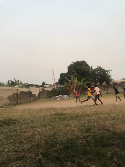 This image, rotated 90 degrees clockwise from its intended upright orientation, depicts an outdoor scene in what appears to be a rural or semi-urban setting. The sky, which is a light, hazy grey, occupies the left side of the frame. An unpaved dirt path runs diagonally from the lower-center towards the upper-left, serving as the main stage for the activity.
Several young males, likely children or teenagers, are visible on or near this path, appearing to be in motion as if running or playing. In the more prominent foreground (lower-mid section when viewed upright), two boys are depicted barefoot or in light footwear: one in a white and orange shirt, and another in a yellow shirt and light blue shorts. Further along the path, near the bottom edge of the frame, another boy in a red shirt is seen next to a pile of colorful discarded items or rubbish. Other figures, less distinct, are visible further up the path.
To the "left" of the path (which would be the background on the left side in an upright view), there are various green trees, including large, leafy specimens and what resembles banana plants. A utility pole with wires extends from the bottom of the frame upwards, piercing the hazy sky. Simple, block-constructed walls, possibly incomplete or damaged, are also present along this side. To the "right" of the path (the foreground on the right side in an upright view), lies an expanse of dry, yellowish-green grass and sparse vegetation.
The time of day is daytime, with an overcast or hazy sky that diffuses the light, resulting in soft illumination and an absence of sharp shadows. The scene portrays a sense of everyday activity in a community area, consistent with the location context of Umudike, Nigeria. There is no visible text in the image.