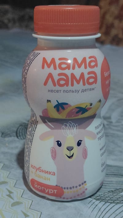 The image shows a bottle of yogurt with a cartoon llama on it. The bottle has a white body with pink and orange accents. The label on the bottle has the brand name "Мама Лама" (Mama Lama) written in large, orange letters. Below that, it says "Несет пользу детям" (Brings benefit to children). There is a picture of a bowl of fruit, including bananas and strawberries, on the bottle. The llama is depicted wearing the bowl of fruit on its head. Text in Russian appears on the bottle, describing the flavor as "Клубника + банан" (Strawberry + banana) and the product as "Йогурт" (Yogurt). Text in a smaller font is also visible on the left side of the bottle. The background is blurred and appears to be a surface with a patterned texture. There is no indication of the city of Ak-Terek, Kyrgyzstan, or any other specific location in the image.