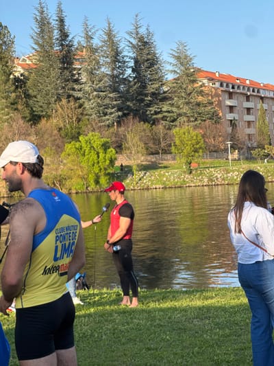 The scene depicts an outdoor interview by a body of water in Mirandela, Portugal, under clear blue skies with strong sunlight. In the mid-ground, a male subject stands barefoot on green grass, facing left, wearing a red baseball cap, a red sleeveless athletic top, and black athletic leggings. He holds a small water bottle in his left hand. An unseen individual extends a microphone with a black foam cover and a white logo, possibly "TVI," towards him for an interview.
To the immediate left of the interviewed subject, partially in the foreground, the back of another male subject is visible. He wears a white baseball cap, a yellow and blue sleeveless athletic vest with the text "CLUBE NÁUTICO PONTE DE LIMA" and "Intermarché," and black athletic shorts. To the right of the interviewed subject, also with her back to the camera, stands a female with long dark hair, dressed in a light-colored long-sleeved collared shirt and blue jeans, with a tan strap across her body.
The background features a calm body of water, likely a river or canal, reflecting the sky and surrounding trees. Beyond the water, the far bank is lined with green grass, various trees including tall conifers and deciduous trees with some bare branches and fresh green foliage. Behind the trees, several multi-story residential buildings with terracotta-tiled roofs are visible, along with a prominent light-brown building featuring balconies. A red park bench and a low fence are also discernible on the far bank. A distant, dark human-like figure is faintly visible near a patch of bright green trees on the far bank.