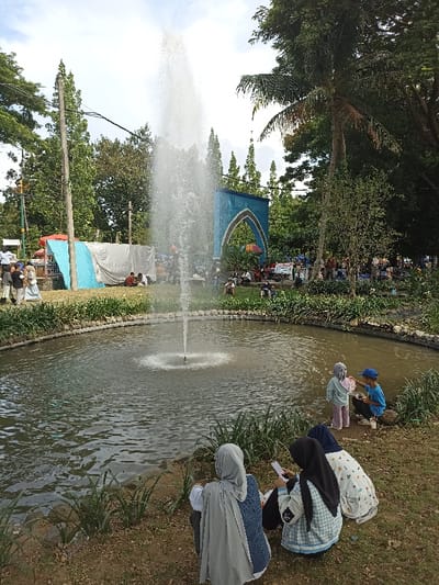 This image captures a vibrant outdoor scene in a public park or garden, likely in Mataram, Indonesia. A circular pond with a central fountain spraying water high into the air dominates the mid-ground. The pond is surrounded by a low stone wall, with grass and sparse vegetation on its banks.
In the foreground, three women, all wearing hijabs, are seated on the grass with their backs to the viewer, facing the pond. One woman in a grey and blue outfit appears to be looking towards the pond, while another in a black hijab and checkered top is holding a piece of paper, possibly reading or writing. A third woman in a white and blue patterned dress is seated beside her. To their right, closer to the pond, two children are visible: one child in a light-colored outfit stands while another in a blue cap squats near the water's edge.
The background is filled with lush greenery, including tall trees and a distinctive blue archway decorated with white patterns, suggesting an architectural feature or entrance. Numerous people are scattered throughout the park behind the pond, sitting on the grass or standing, indicating a public gathering or recreational day. To the left of the archway, temporary structures or market stalls, covered with blue and white tarpaulins, are set up, implying a market or community event is taking place.
The time of day appears to be daytime, possibly morning or early afternoon, under a bright sky with scattered clouds, and the weather seems pleasant. The overall atmosphere is relaxed and social. A white sign with dark text is visible near the blue archway, but the text is too small to be deciphered.