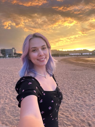 A young woman with dyed purple and pink hair smiles at the camera while taking a selfie on a sandy beach. She is wearing a black dress with a floral print and a pearl necklace with a small sun pendant. The beach is mostly empty, with buildings and hills visible in the background. The sky is filled with dramatic orange and yellow clouds, indicating either a sunset or sunrise. The location is the Playa de Poniente in Gijón/Xixón, Spain.