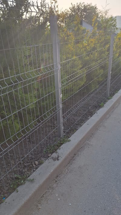 The image depicts a wire mesh fence with vertical posts, bordering a paved area and a patch of vegetation. The fence is constructed from interconnected metal wires, forming a grid pattern. The posts are metallic and appear to be gray. Behind the fence, dense green foliage is visible, likely shrubs or trees. A concrete curb separates the paved surface from the area enclosed by the fence. The paved surface appears to be asphalt and shows some debris and tire marks. The environment suggests an outdoor setting, possibly a street or pathway adjacent to a property. The photograph was taken in Sărata-Galbenă, Moldova. There are no people or animals visible in the image. The lighting suggests it is daytime, possibly late afternoon given the angle of the sunlight.