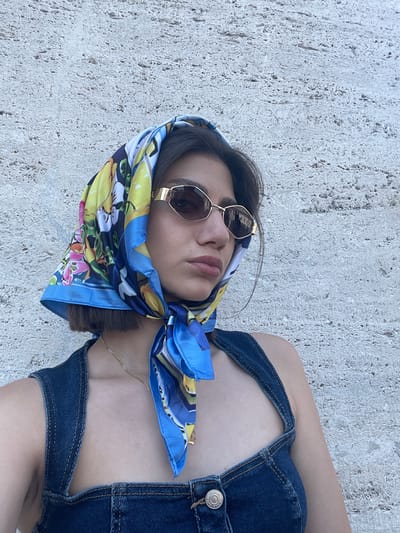 A woman is lying on a textured, light gray surface, possibly stone or concrete. She wears a colorful headscarf with blue, yellow, and floral patterns, as well as gold-rimmed sunglasses. Her clothing appears to be denim overalls, and a gold chain necklace is visible around her neck. The location is Città del Vaticano, Vatican City.