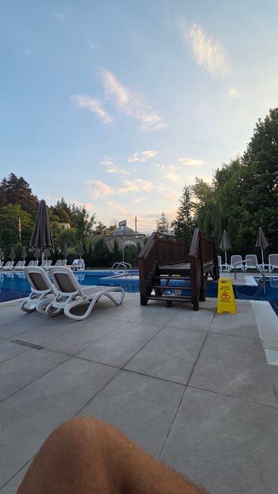 The image captures a poolside scene at dusk in Bania, Bulgaria. The sky is a soft blue with scattered, wispy clouds tinged with orange from the setting sun. Trees line the background, with a building structure visible amongst them. In the foreground, the tiled poolside area features several lounge chairs, some stacked and others open, with umbrellas providing shade. A wooden structure with steps and seating is situated next to the pool. A bright yellow "CAUTION WET FLOOR" sign with a warning symbol is placed near the pool's edge. The water in the pool is a vibrant blue. The overall impression is one of a peaceful, end-of-day setting at a resort or hotel. The perspective suggests the photo was taken by someone standing near the pool, possibly about to enter or having just left the water, as a portion of an arm is visible in the lower right corner.