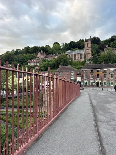 The image shows a view from a bridge in Ironbridge, United Kingdom, looking towards the town situated on a hillside. A rust-colored metal fence runs along the left side of the bridge. The road surface is asphalt, with a metal strip running down its center.

In the background, a row of brick buildings with green shutters lines the street. One building has a sign that reads "THE SEVERN HOUSE HOTEL." A church with a prominent clock tower stands on the hilltop, surrounded by lush green trees. Houses are scattered across the hillside, partially obscured by foliage. The sky is overcast, with a muted light. A single person, dressed in dark clothing, is walking away from the camera on the right side of the bridge. Below the bridge and visible through the fence, a garden area with planters and benches can be seen.