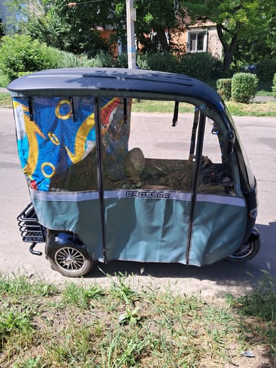 An outdoor, eye-level, medium shot captures a dark gray three-wheeled electric vehicle parked on an asphalt road. The vehicle is covered with a transparent plastic enclosure, revealing a patterned fabric interior with gold rings on a blue background. The vehicle has a black metal rack at the rear and the word "GROSSER" is visible in silver lettering on the side. The scene is bordered by a grassy verge on the right and left, with green trees and a building visible in the background. The lighting suggests it is daytime, likely in the city of Kamianske, Ukraine.