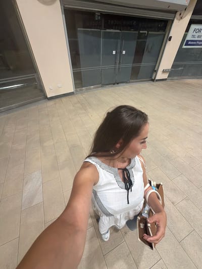 A young woman is captured in a selfie taken at a slightly downward angle, standing on a tiled walkway. She has long, dark hair and is wearing a white dress with black trim and a bow at the neckline. She is also carrying a brown woven bag and wearing white sneakers. The background shows the exterior of a building with large glass doors and a beige facade. A sign with text that is partially obscured is visible on the upper left. The lighting suggests it might be daytime, possibly evening. The setting appears to be an outdoor area, possibly a commercial or public space, in Nicosia, Cyprus. The woman's expression is neutral, and she is looking slightly away from the camera.