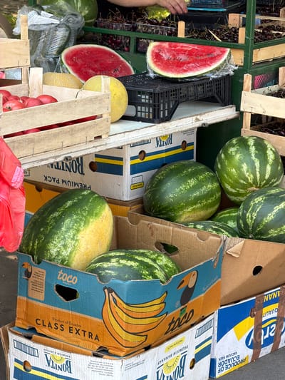 This image depicts a fruit stand in Palilula Urban Municipality, Serbia, filled with various fruits for sale. In the foreground, several large watermelons are displayed, some in cardboard boxes. One box, labeled "toco BANANAS," features an illustration of bananas and a toucan. Adjacent to the watermelons, a wooden crate holds red tomatoes. Further back, a halved watermelon is visible, wrapped in plastic. Melons are also present in a wooden crate. In the background, shelves are stacked with containers of cherries. The overall scene suggests an outdoor market setting.