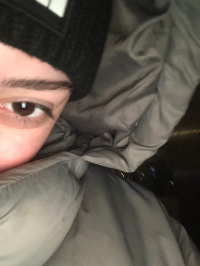 A close-up shot shows a person wearing a black hat, nestled between the folds of a gray puffy jacket. The focus is primarily on one eye, which is brown. The jacket seems to envelop the person, with the fabric bunched around their head and shoulders. The background is out of focus, hinting at darkness and possibly a nighttime setting.