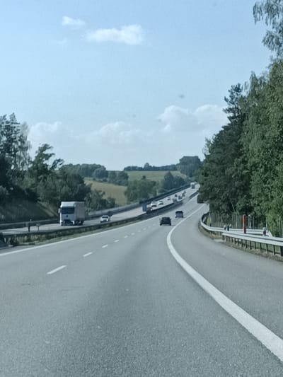 The image depicts a highway scene in Ostředek, Czechia, under a bright, partly cloudy sky, suggesting daytime. The camera is positioned from a moving vehicle, looking forward along a multi-lane highway that curves to the right. Dense green trees line the right side of the road, with a slightly hilly, cultivated landscape visible in the distance on the left.
Several vehicles are present on the highway. A silver truck is in the left lane of the highway, and a white car is in the lane to its right. Further ahead, traffic is visible, including a dark car in the left lane and other vehicles in the distance. The road markings are clear, with solid white lines separating lanes and dashed white lines indicating the path of travel. A metal guardrail runs along the right side of the highway, punctuated by red and white markers. The overall impression is one of typical highway travel on a clear day. No text is visible in the image.