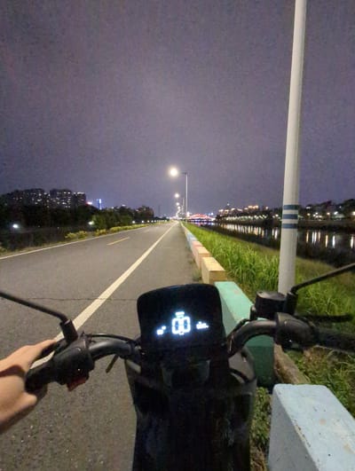 A person is riding a scooter or bicycle on a paved path with a white line marking the edge. Streetlights illuminate the path, which stretches into the distance. On the left side of the path, there are buildings and trees. On the right side, there is grass and a body of water reflecting lights from the city. The scene appears to be at night, in Shunde District, China. The scooter or bicycle's dashboard displays information, possibly including speed. The person's hand is visible near the scooter's controls.