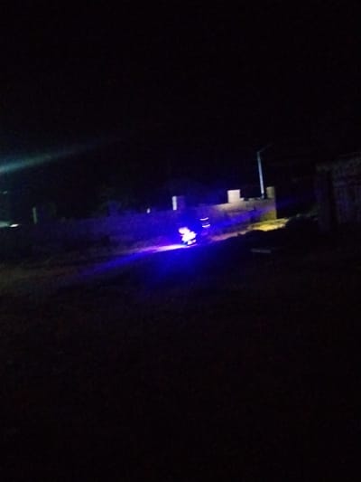 The image shows a dark, outdoor scene, likely at night, with some sources of light. In the lower center of the image, a bright blue light emanates from what appears to be a vehicle, possibly a motorcycle or a scooter, with some reflective elements visible. The light casts an illumination on the dark ground and surrounding structures. The background is largely obscured by darkness, but hints of buildings and a lamppost are discernible. The overall impression is that of a dimly lit urban or semi-urban environment.