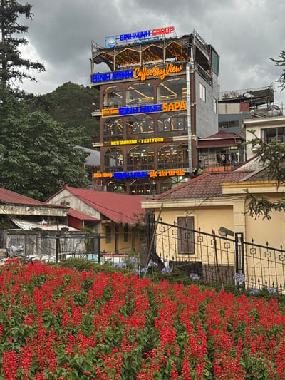 The image captures a street view in the town of Sa Pa Ward, Vietnam, focused on a tall building with the sign "BINH MINH Coffee Sky View NINH SAPA RESTAURANT FAST FOOD ĐẶC SẢN TÂY BẮC" displayed in bright blue and orange neon lettering. The building appears to be a restaurant or cafe, with multiple floors and large windows. There are also red flowers lining the foreground, bordering a wrought iron fence in front of a light yellow building. In the background, trees and other buildings are visible. The sky is overcast, suggesting a cloudy day.