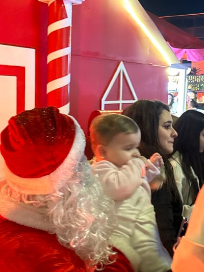A baby in light pink attire sits with a woman near a Santa Claus figure, complete with a red hat and white beard. The backdrop includes a red structure with white candy cane-striped poles and a white triangular design at the top. The image has a slightly tilted orientation.