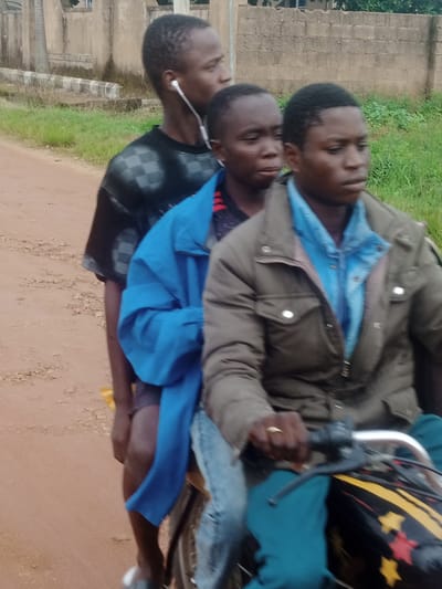 The media file shows three young males riding on a motorcycle. The person in the front is wearing a dark brown jacket over a blue shirt and appears to be driving. The person in the middle is wearing a bright blue jacket and a dark shirt underneath. The person in the back is wearing a black t-shirt with a checkered pattern and has white earphones in. All three individuals appear to be of African descent.
The scene is outdoors, with a dirt road and a grassy area visible. In the background, there is a beige building with some greenery. The lighting suggests it is daytime, possibly overcast. The individuals are positioned as if they are in motion, with the focus on their upper bodies and the motorcycle. The overall impression is that of a casual journey or commute. There are no visible signs of text or specific branding. The context provided indicates the location is Katon Rikkos, Nigeria.