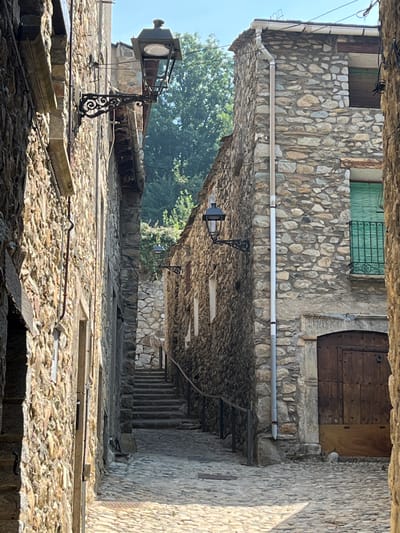The image shows a narrow, cobblestone alleyway in Benasque, Spain, flanked by old stone buildings. The buildings have rough-hewn stone walls and some feature traditional architectural elements like wrought-iron lampposts and wooden doors. In the distance, a stone staircase ascends between the buildings, leading towards more stone structures and greenery. The sky is clear and blue, suggesting a sunny day. There are no people visible in the image. The scene is one of an old, rustic European town.