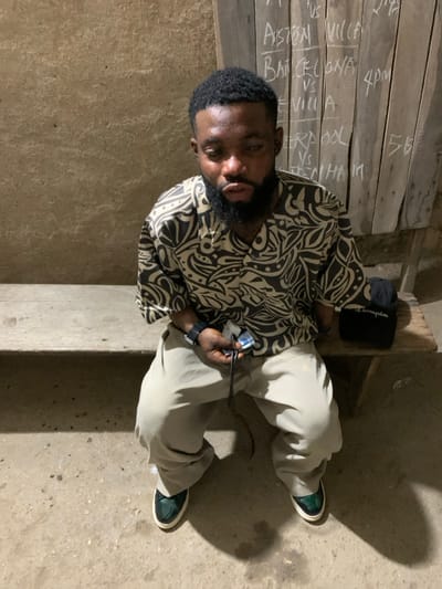 The image displays an adult male seated on a wooden bench in an establishment located in Okene, Nigeria. The individual has dark skin, a full beard, and short, dark hair. He is wearing a short-sleeved, collared shirt featuring a black and beige abstract pattern, light-colored trousers, and dark green shoes with white soles. A dark wristwatch is visible on his left wrist. In his left hand, he holds a small, light-colored object and a black cord. To his right, on the bench, rests a dark object with "Champion" written in white. The background on the left consists of a textured, light brown wall. To the right, a vertical wooden board, assembled from multiple planks, functions as a chalkboard. Handwritten in white chalk on this board are several football (soccer) match listings, including "ASTON VILLA VS", "BARCELONA VS", "SEVILLA", "LIVERPOOL VS", and "TOTTENHAM", along with times such as "4pm" and "56". The floor is a rough, light-colored concrete or packed earth surface. The scene is illuminated by an overhead light source, casting distinct shadows.