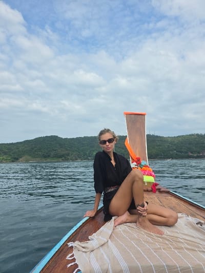 A slender woman in a black cover-up and sunglasses sits on the front of a longtail boat, her legs tucked. A striped towel is draped beside her. The boat's wooden structure is visible, with colorful decorations adorning the prow. The water is calm with a forested coastline in the background. Cloudy skies fill the top left of the frame. The image is oriented vertically.