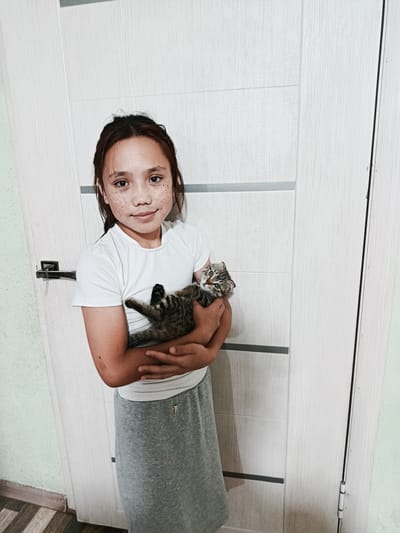 A young girl with freckles stands in front of a light-colored door with horizontal paneling. She is wearing a white t-shirt and a grey skirt. She is holding a small, tabby kitten in her arms. The kitten is facing the camera and looking towards the viewer. The door appears to be an interior door with a modern design. The background wall is a pale green color. The flooring is a wood-grain pattern. The lighting suggests an indoor setting, possibly a home. The image quality is slightly grainy, indicating it may have been taken with a mobile device or in low light conditions. The overall scene depicts a candid moment between a child and a pet.