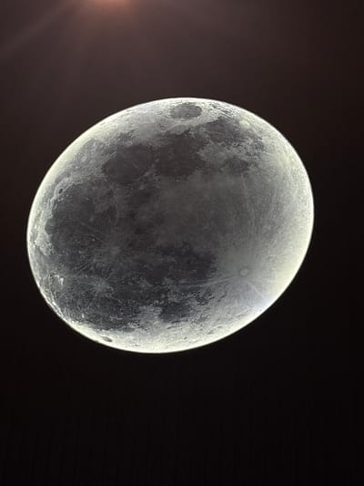 The image shows a close-up, detailed view of the moon against a dark background. The moon's surface is clearly visible, with craters and maria (dark, smooth plains) easily discernible. The lighting suggests the moon is illuminated from the side, creating a crescent shape with a bright rim and detailed shadows on the unlit portion. The background is uniformly dark, providing a stark contrast that emphasizes the moon's texture and features. There are no people, buildings, or any indication of a specific location within the city of Orlando, United States of America, as the image is focused solely on the celestial body. The top portion of the image contains some faint light rays, possibly indicative of a light source or atmospheric effect near the top edge.