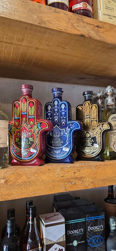 The image shows a close-up, vertical view of several bottles of liquor displayed on wooden shelves. The bottles are diverse in shape and color, with some featuring intricate hand-shaped designs and others being more traditional in form. The hand-shaped bottles, in gold, blue, and red, appear to be a type of tequila. Their decorative patterns are prominent. Among the other bottles, some are clearly labeled as Jack Daniel's Tennessee Honey. Further to the right, there are boxes labeled "DOORLY'S BARBADOS RUM 12 YEARS". The overall setting suggests a liquor store or a well-stocked bar. The lighting is ambient, typical of an indoor retail environment. No people are visible in the image. The wooden shelves have a rustic appearance. Visible text includes brand names like "JACK DANIEL'S", "DOORLY'S", and "CASA DE HAMSA", as well as descriptions such as "Tennessee Honey", "BARBADOS RUM", "AGED 12 YEARS", "TEQUILA BLANCO", and "TEQUILA ANEJO". The location context is stated as Jerusalem, Israel, which is not directly inferable from the image itself.