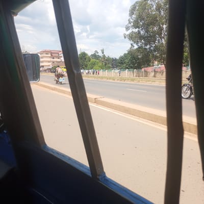 The scene depicts a daytime street view in Sheywe ward, Kenya, observed from inside a vehicle through its interior structural bars, which are dark blue and metallic. A multi-lane asphalt road with a concrete median strip runs horizontally across the mid-ground. On the left lane, a male individual operates a motorcycle, which is heavily laden with multiple large, light-colored sacks secured to the rear. The motorcycle's license plate or rear sign is partially visible, reading "KING DAVID". Further down the road, a yellow vehicle is partially visible among other traffic.
On the right side of the road, separated by the concrete median, a sidewalk runs parallel to the street. A group of approximately 10-15 pedestrians, dressed in various casual attire including white shirts, dark trousers, and some with backpacks, are walking in a line along this sidewalk. They are positioned behind a low, light-colored brick wall topped with a white and light blue ornamental metal fence. Beyond the fence, dense green foliage from numerous trees is visible, with a large, mature tree dominating the upper right background.
In the left background, behind the motorcyclist and across the road, a multi-story, light-colored building with a "GUEST HOUSE" sign on its upper facade is prominent. Other smaller buildings and structures with reddish-brown roofs and visible solar water heaters are interspersed within the green landscape. The sky is partially cloudy with patches of blue. Another motorcycle is partially visible on the far right of the frame, near the median. The road surface appears dry and clear.