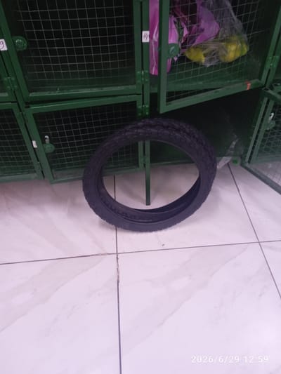 A black bicycle tire lies on a tiled floor in front of a row of green metal lockers. The lockers have wire mesh doors. Some of the lockers contain items, including a bag with yellow objects, possibly fruit, in one locker. The floor tiles are white with grey veining. The timestamp "2026/6/29 12:59" is visible in the lower right corner. There is no indication of the location being in Ak-Suu, Kyrgyzstan.
