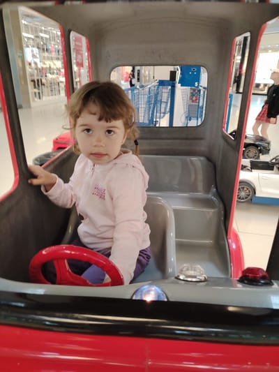 A young girl with brown hair and pigtails sits in the driver's seat of a red toy car. She is wearing a pink zip-up hoodie with a graphic on the front and purple pants. Her left hand rests on the red steering wheel. The toy car has a gray interior with seats and a dashboard. In the background, blurry shopping carts are visible, suggesting the setting is a mall or a large store. To the right, other toy cars, including a black one and a white one, are parked. The girl appears to be looking towards the viewer with a neutral expression. The lighting suggests it is daytime. The location is Wołomin, Poland.