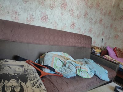 The image depicts an indoor scene, likely a bedroom or living area, with a sofa and a wall covered in floral wallpaper. On the sofa, there are several items including a rumpled blue blanket with a geometric pattern, a pillow with a black and white comic strip design, and an orange plastic clothes hanger. To the right of the sofa, another bed is visible, also covered with blankets and pillows. The overall impression is one of a lived-in, possibly untidy space. The location is not discernible from the image itself.