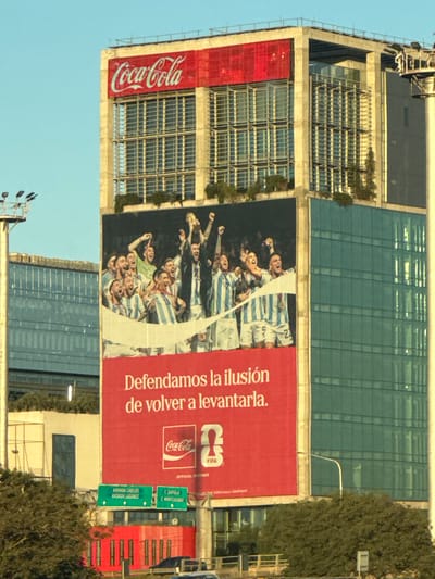 The image captures a large Coca-Cola advertising billboard affixed to the side of a tall building in Vicente López, Argentina. The advertisement features a graphic of the Argentinian national football team celebrating, with the text "Defendamos la ilusión de volver a levantarla" (Let's defend the illusion of rising again). The Coca-Cola logo is prominently displayed, along with the FIFA logo. The background shows a mix of modern architecture and greenery, typical of an urban environment. Street signs are visible indicating nearby locations. The sky is clear, suggesting daytime.