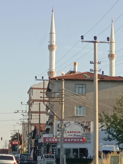The image depicts a street scene in Bahşılı Belediyesi, Turkey, bathed in the warm light of either late afternoon or early morning. The sky is a clear, pale blue, suggesting fair weather. Dominating the left side of the frame are two tall, slender minarets of a mosque, their white spires reaching towards the sky, adorned with intricate decorative bands.
In front of the mosque and to its right, a multi-story building lines the street, its architecture a mix of traditional and modern elements. The building has a reddish-brown tiled roof, and several windows are visible, some with dark frames. Utility poles with numerous electrical wires crisscross the scene, creating a sense of urban infrastructure.
Below the mosque and the building, a commercial street unfolds. Several businesses are visible, indicated by signage. One prominent sign features the word "CELEB" in large red letters, suggesting a retail establishment. Another sign displays "ASPAVA" within a circular logo, and below it, the word "ASPAVA" is repeated in red letters on a rectangular sign. Further down the street, a red sign with the "BİM" logo is visible, indicating a discount supermarket chain.
The street is populated with vehicles, including a white pickup truck with the word "ISUZU" on its tailgate, several cars, and potentially a dark-colored van. The presence of these vehicles suggests that the street is active with traffic. The overall impression is a bustling, everyday scene in a Turkish town, with religious structures coexisting alongside commercial activity and daily life. No specific emotional tone is conveyed; the image is a straightforward visual documentation of the location.
