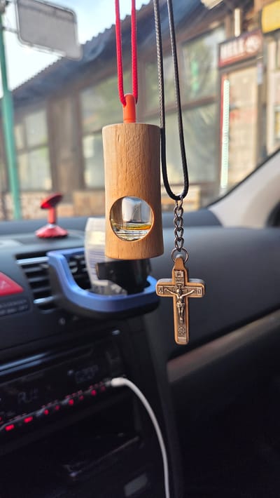 In Razlog, Bulgaria, a close-up shot captures a wooden car air freshener with a small glass vial of liquid hanging from the rearview mirror. Below the air freshener, a metal Christian cross hangs on a chain. The car's dashboard is visible, including a car radio with illuminated red buttons and a white cable plugged into it. In the background, the exterior of a building with windows and a corrugated roof is seen, suggesting an urban setting.